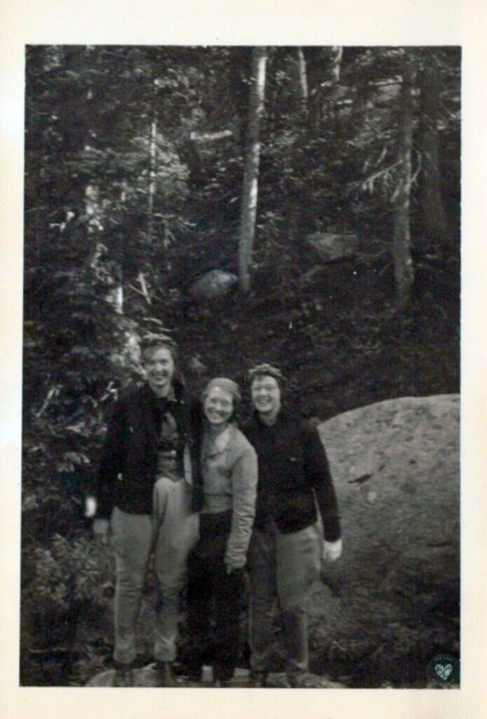 Black and white photo of three women standing outside in a forest on boulders
